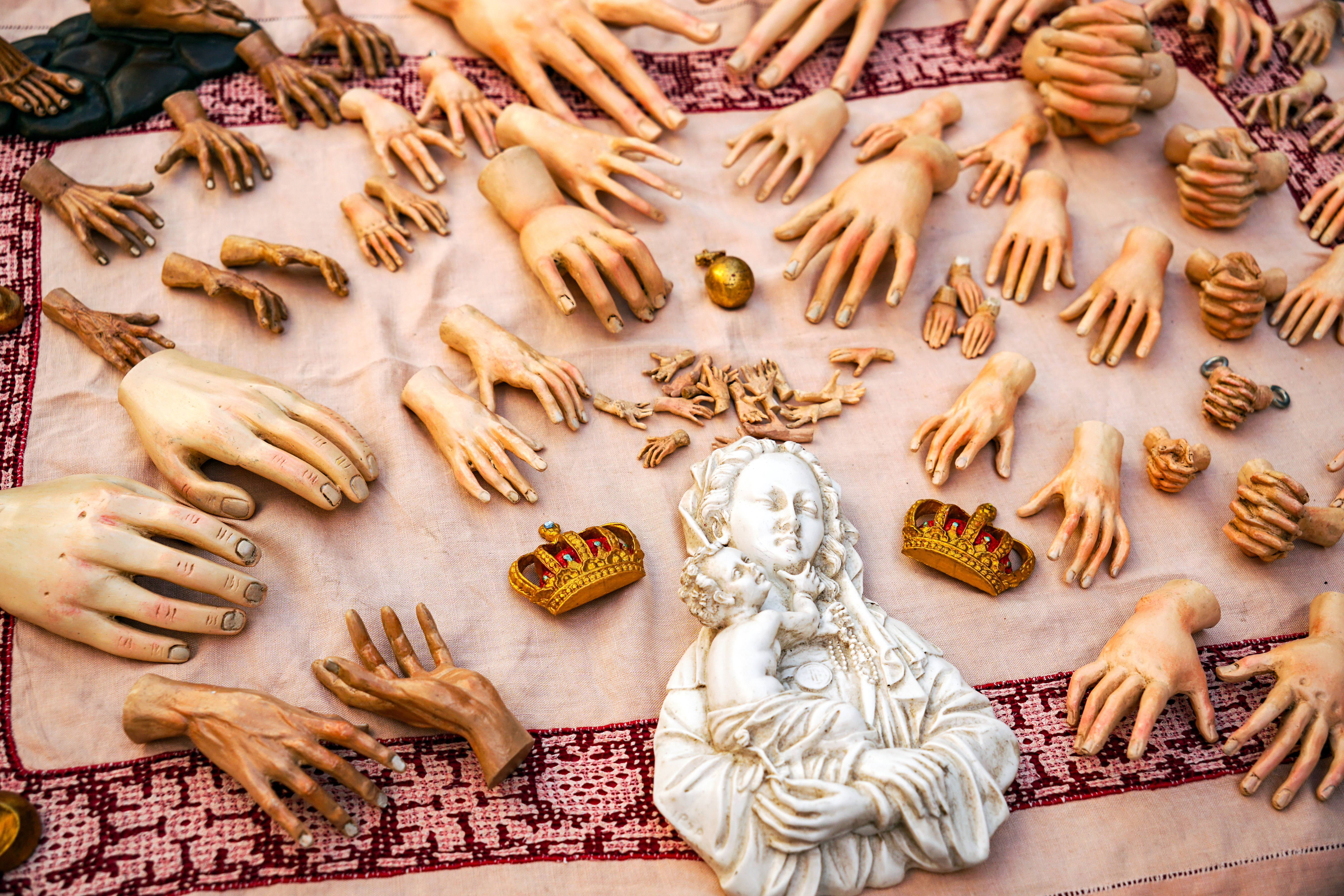 A variety of hand sculptures and figures are arranged on a light fabric surface, with a focus on both large and small hands in different poses and styles. Among them is a white figurine of a mother holding a child, flanked by two small golden crowns.