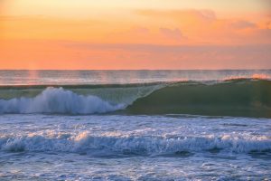 
A serene ocean scene at sunrise featuring a gently curling wave illuminated by soft orange and pink hues in the sky. The water is frothy with whitecaps, and the horizon blends quietly into the colorful sky.