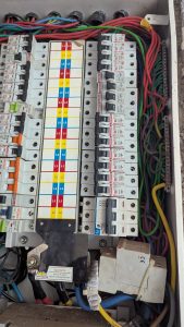 A close-up view of an electrical distribution board, featuring multiple circuit breakers with labeled switches, organized in rows. Color-coded wires in red, green, blue, yellow, and black are visible, leading into the board.