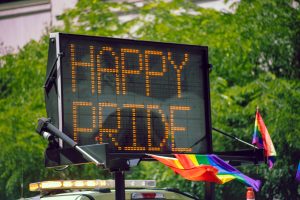 A digital road sign displays the message "HAPPY PRIDE" in orange lights during a Pride celebration. Rainbow flags are attached to the vehicle below the sign, which is parked in front of a backdrop of lush green trees.