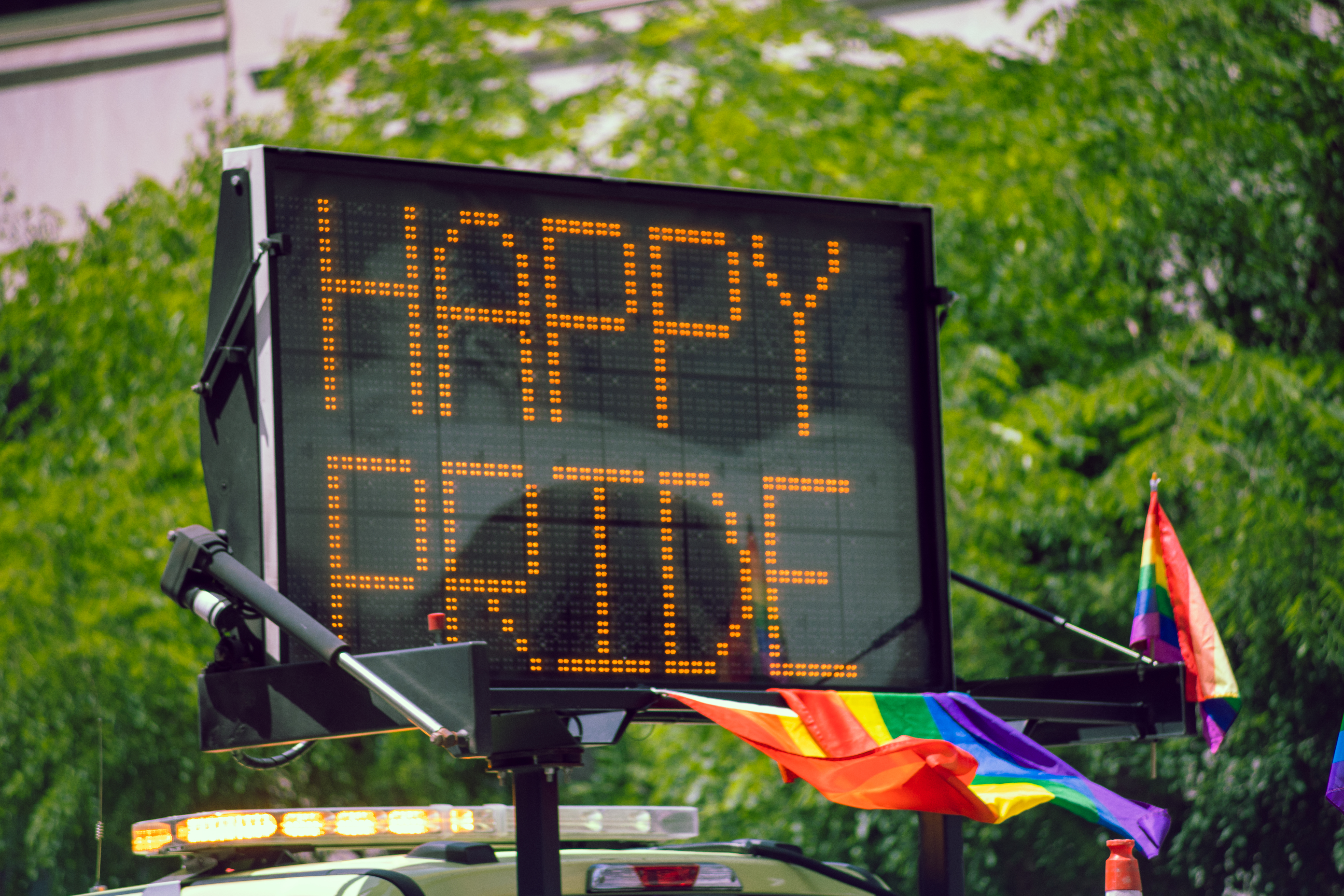 A digital road sign displays the message "HAPPY PRIDE" in orange lights during a Pride celebration. Rainbow flags are attached to the vehicle below the sign, which is parked in front of a backdrop of lush green trees.