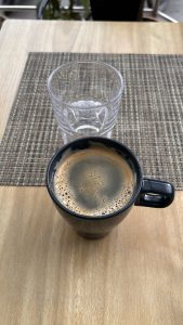 A top-down view of a black coffee mug filled with dark coffee, featuring a frothy surface. Next to the mug is a clear glass tumbler that is empty.