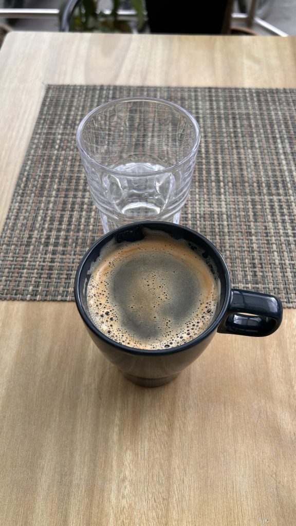 A top-down view of a black coffee mug filled with dark coffee, featuring a frothy surface. Next to the mug is a clear glass tumbler that is empty.