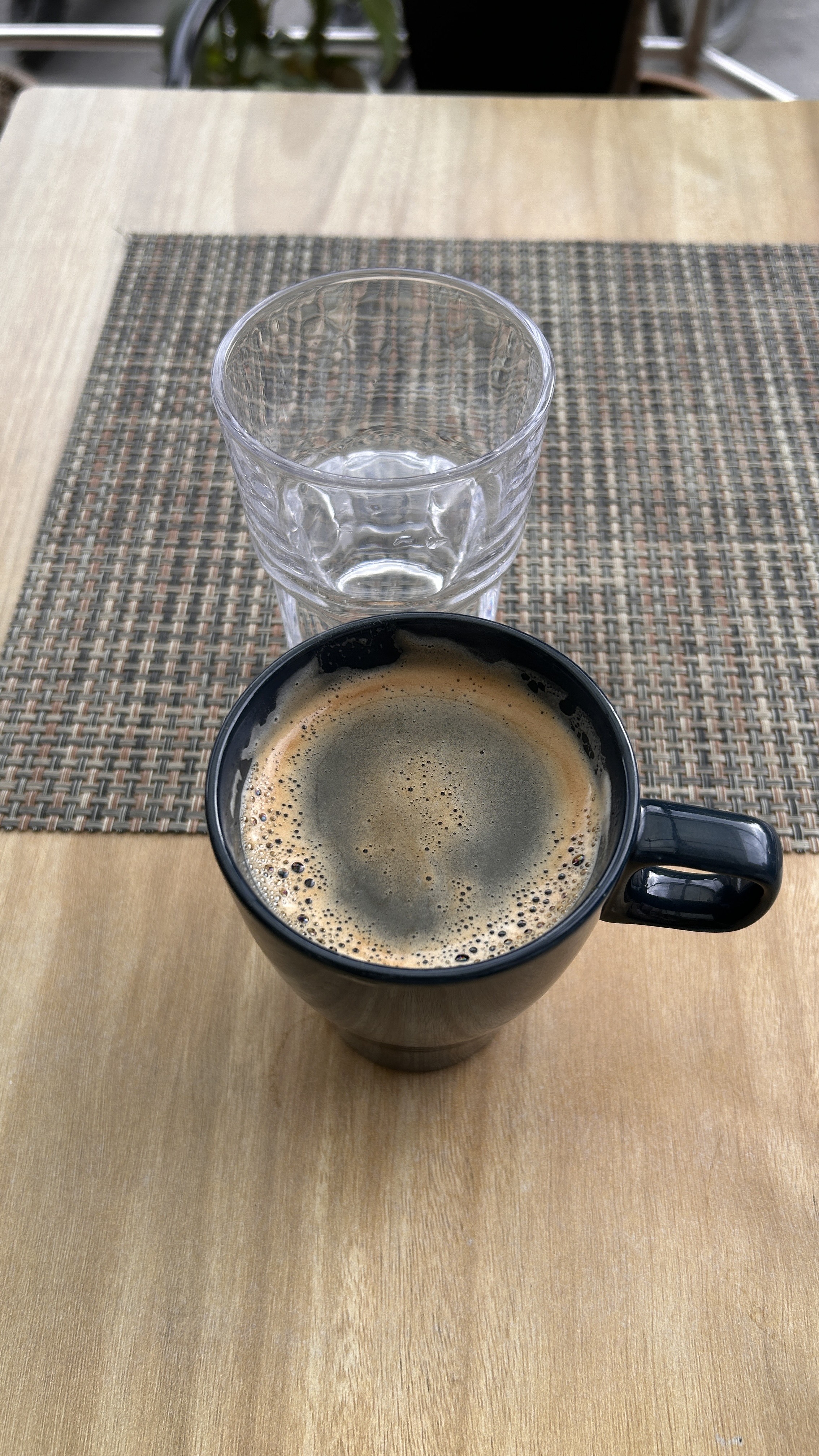 A top-down view of a black coffee mug filled with dark coffee, featuring a frothy surface. Next to the mug is a clear glass tumbler that is empty.