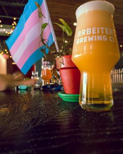 A Beer sits on a wood bar with a flower pot in the background. The flower pot has a small green plant and a trans pride flag.