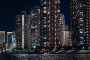 A night view of Tokyo showing the Sumida River and the high-rise apartment buildings towering along it. A Yakatabune(Japanese houseboat) with a glowing red lantern slowly sails past the apartment buildings.
