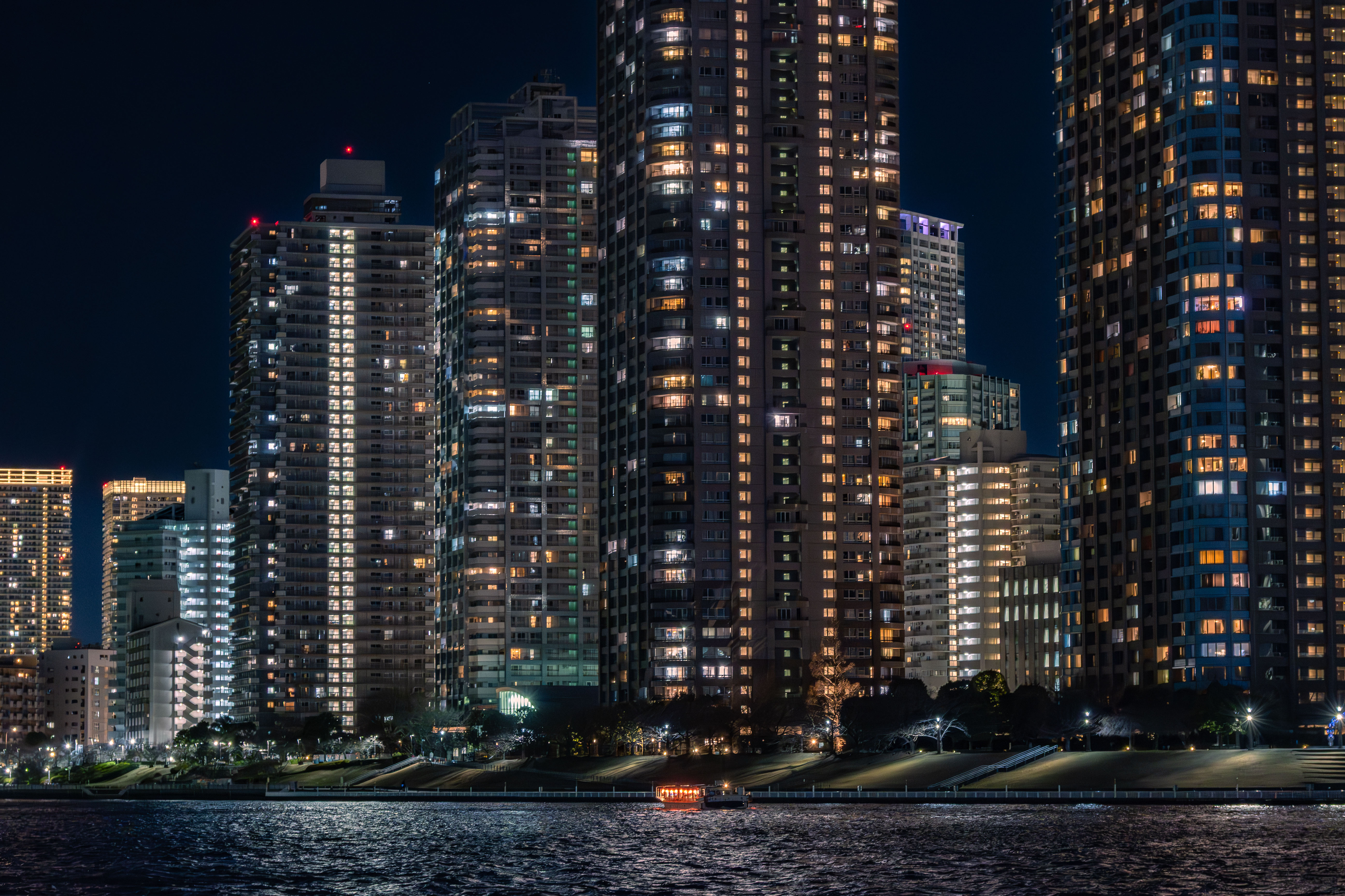A night view of Tokyo showing the Sumida River and the high-rise apartment buildings towering along it. A Yakatabune(Japanese houseboat) with a glowing red lantern slowly sails past the apartment buildings.