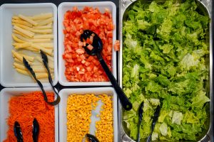 The image shows a salad bar setup with various ingredients in white and silver containers. There are sliced asparagus, diced tomatoes, shredded carrots, and corn kernels right container. Fresh green lettuce is displayed in a larger silver tray.