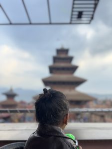 A young child with dark hair is seen from behind, overlooking a traditional multi-tiered pagoda style building in the background. 
