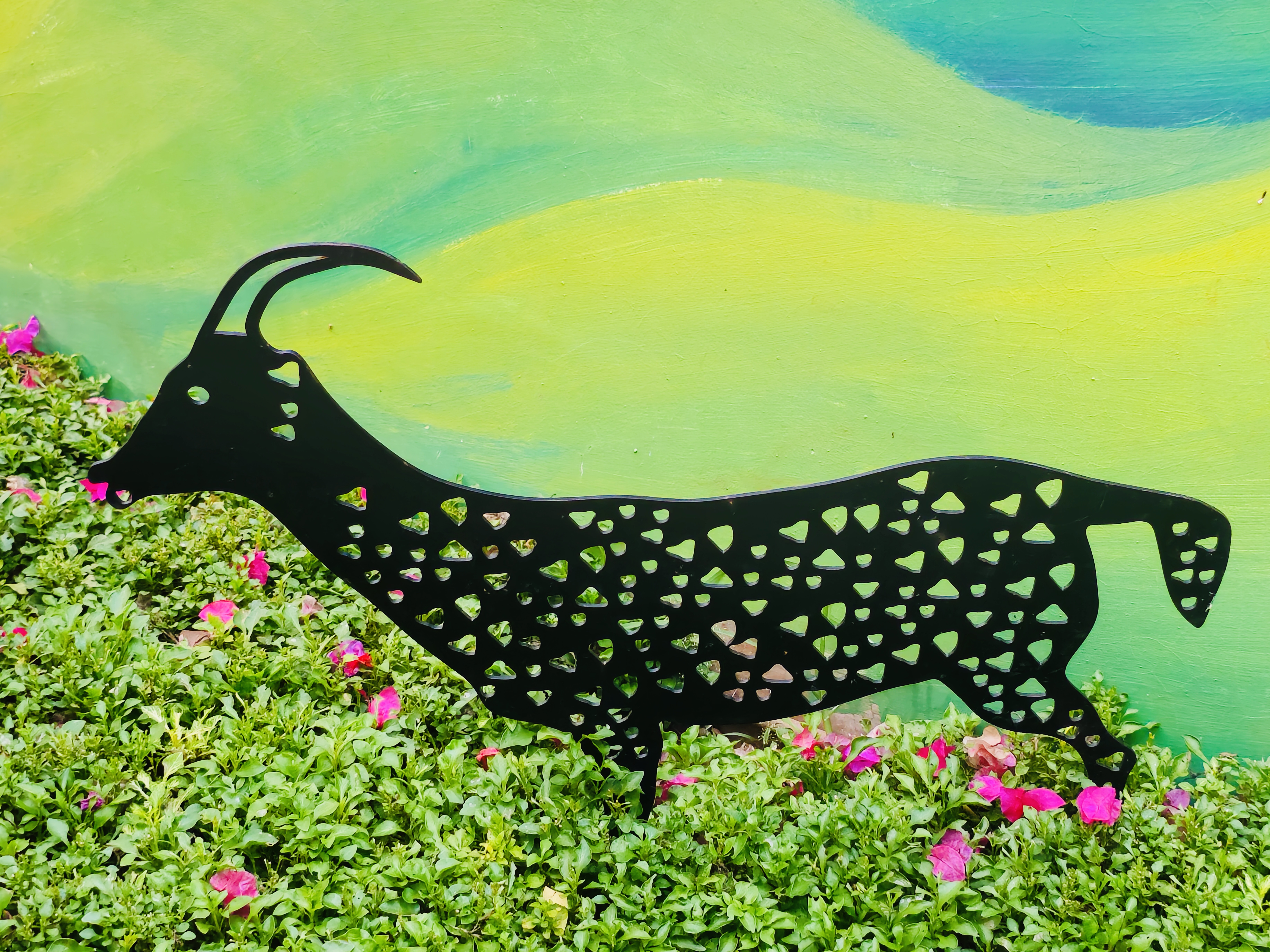 A black metal goat silhouette with heart cutouts nestled in a green bush with pink flowers. Captured in Kuttiady, Kozhikode.
