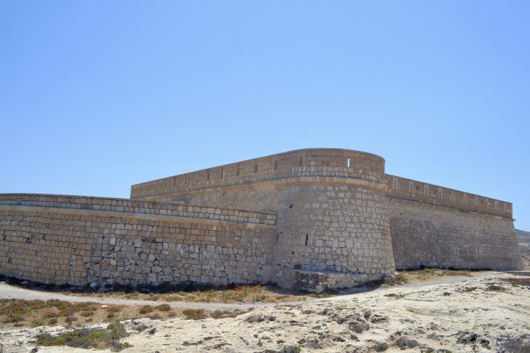 The historic Castillo de San Felipe, a coastal fortress in Los Escullos, Almería, Spain. It has strong stone walls with battlements and stands under a clear blue sky.