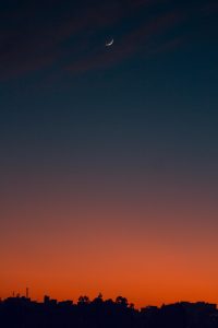 A serene twilight sky displays a gradient of deep blue transitioning to warm orange hues near the horizon. A thin crescent moon is visible in the upper right section of the image, subtly illuminated against the evening sky.