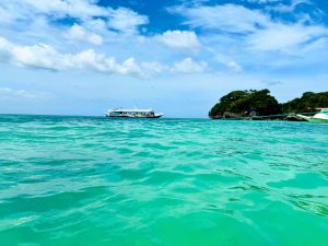 A serene view of clear turquoise waters with gentle ripples, a boat in the distance surrounded by a bright sky scattered with fluffy clouds
