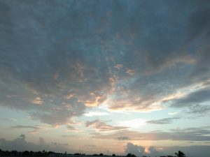 A dramatic sky at sunset, featuring a mix of soft clouds in varying shades of gray and blue, with hints of orange and pink illuminating the lower part of the clouds.