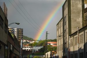 A rainbow arcs over an industrial landscape, emerging from dark rain clouds. The scene captures a view between two buildings, revealing a row of residential apartments in the distance, surrounded by greenery.