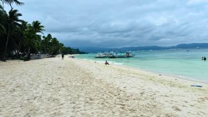 A serene beach scene featuring soft, golden sand and a light turquoise sea under a cloudy sky.
