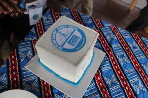 A cake built in the shape of a cube, white frosting with pale ble trim. A label on the top says "WordCamp Jinja 2025"