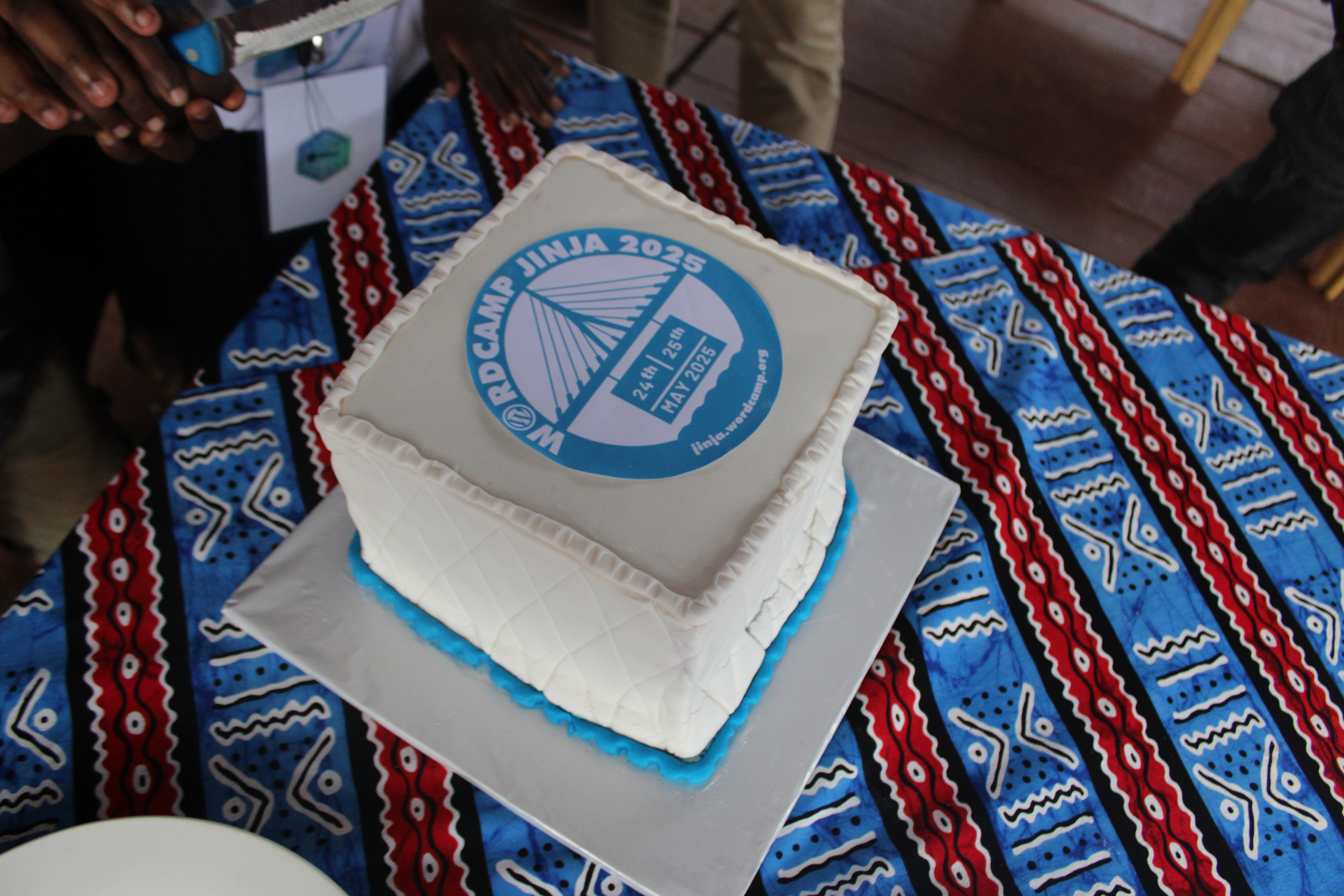 A cake built in the shape of a cube, white frosting with pale ble trim. A label on the top says "WordCamp Jinja 2025"