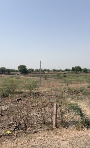A vast expanse of dry land is shown, featuring scattered shrubs and bushes. In the background, a few trees can be seen, along with a small body of water reflecting the clear blue sky.
