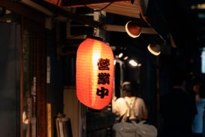 A lantern saying "Open" in a corner of the Marunouchi drinking district in Tokyo. The red lantern is lit up, and behind it you can see the backs of customers drinking.
