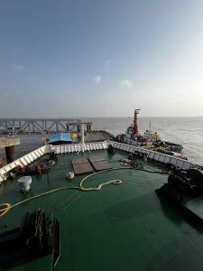 A view from the deck of a ship looking out toward the sea, with a calm horizon in the background. The foreground features a green deck with ropes and equipment, including winches. In the distance, several tugboats are docked near a pier.