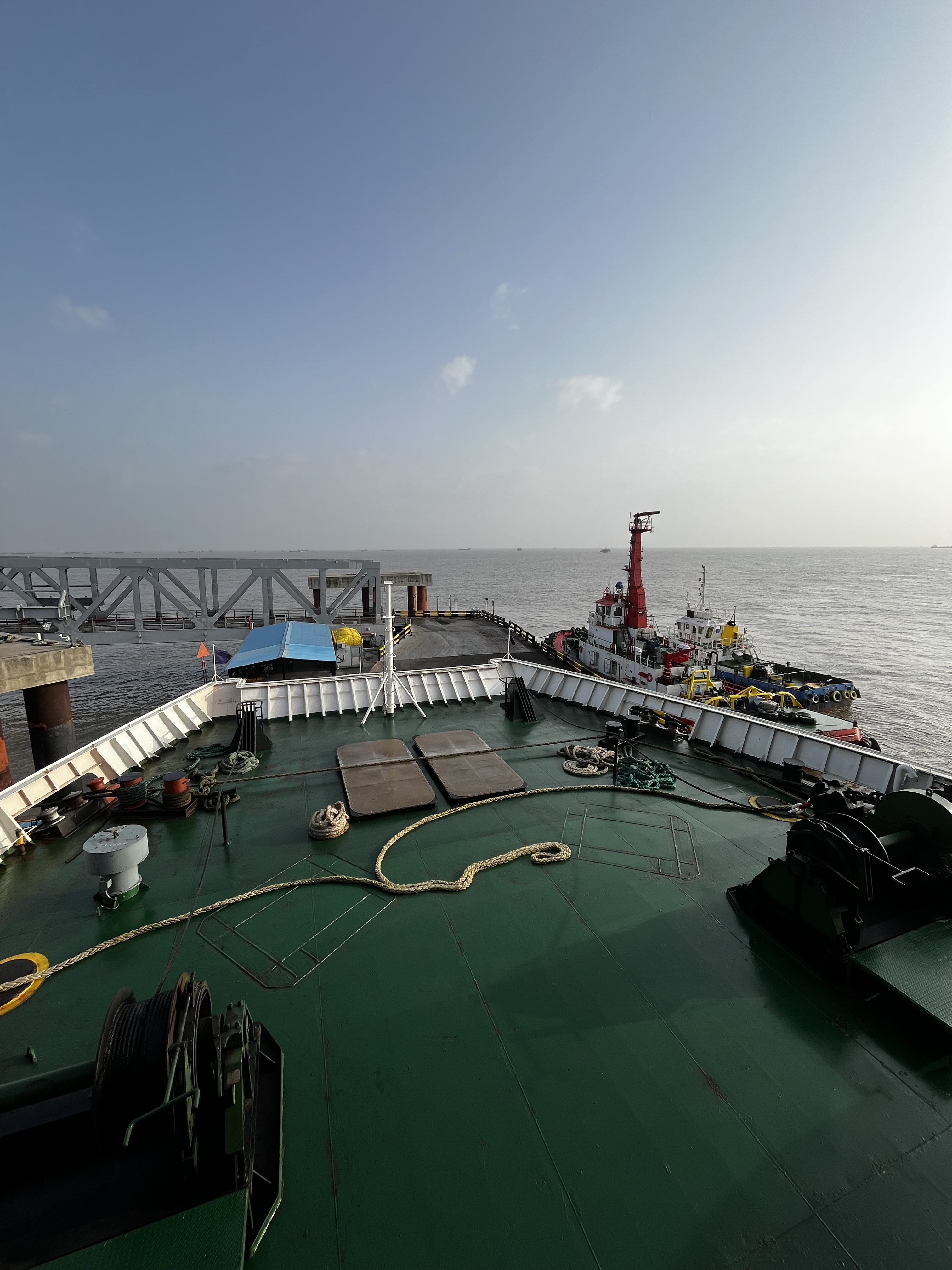 A view from the deck of a ship looking out toward the sea, with a calm horizon in the background. The foreground features a green deck with ropes and equipment, including winches. In the distance, several tugboats are docked near a pier.