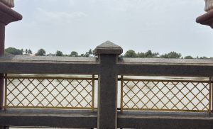 A close-up view of a decorative riverside railing with a crisscross metal design painted in gold. The railing is made of dark stone with a central pillar topped with a pyramid-shaped cap. 