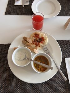 A white plate featuring a portion of dosa topped with chopped tomatoes, accompanied by a serving of sambar (a lentil-based vegetable stew) and a bowl of yogurt.