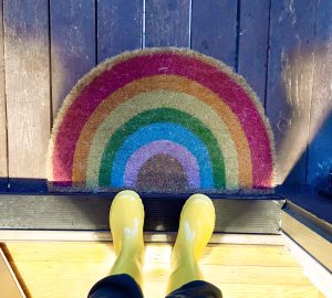 Looking down at someone in dark pants and yellow rain boots standing in an open doorway looking down at a curved rainbow door mat.