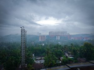 A panoramic view of a misty landscape under a cloudy sky, featuring a tall chimney in the foreground. In the background, there are tall buildings nestled among lush greenery, with a mix of residential structures and trees visible.