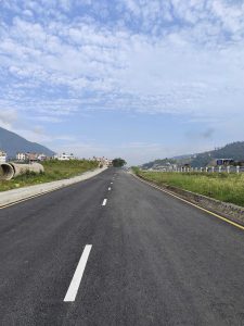 A straight, newly paved road stretches into the distance, flanked by grassy areas and a low concrete barrier. 
