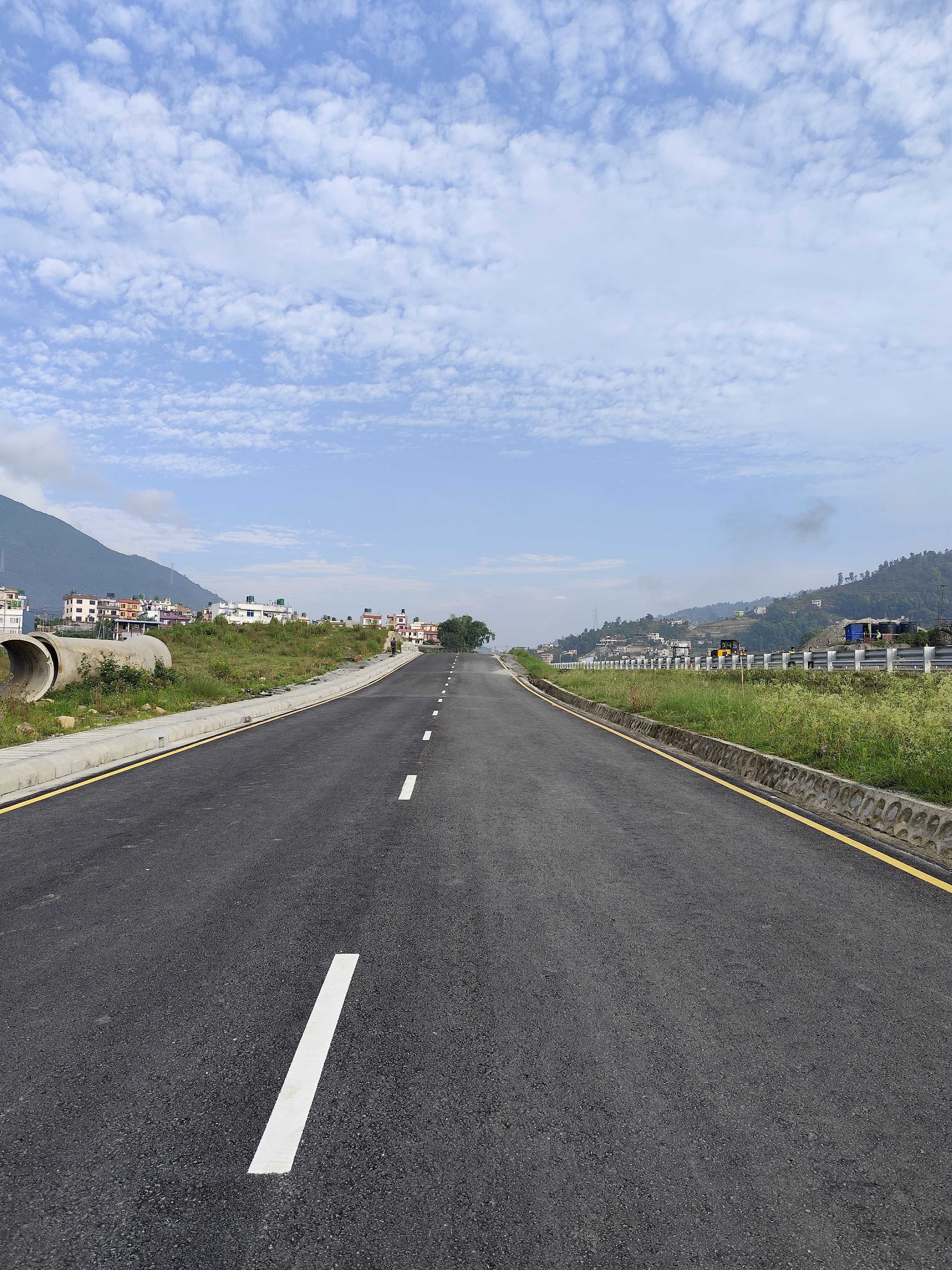 A straight, newly paved road stretches into the distance, flanked by grassy areas and a low concrete barrier.