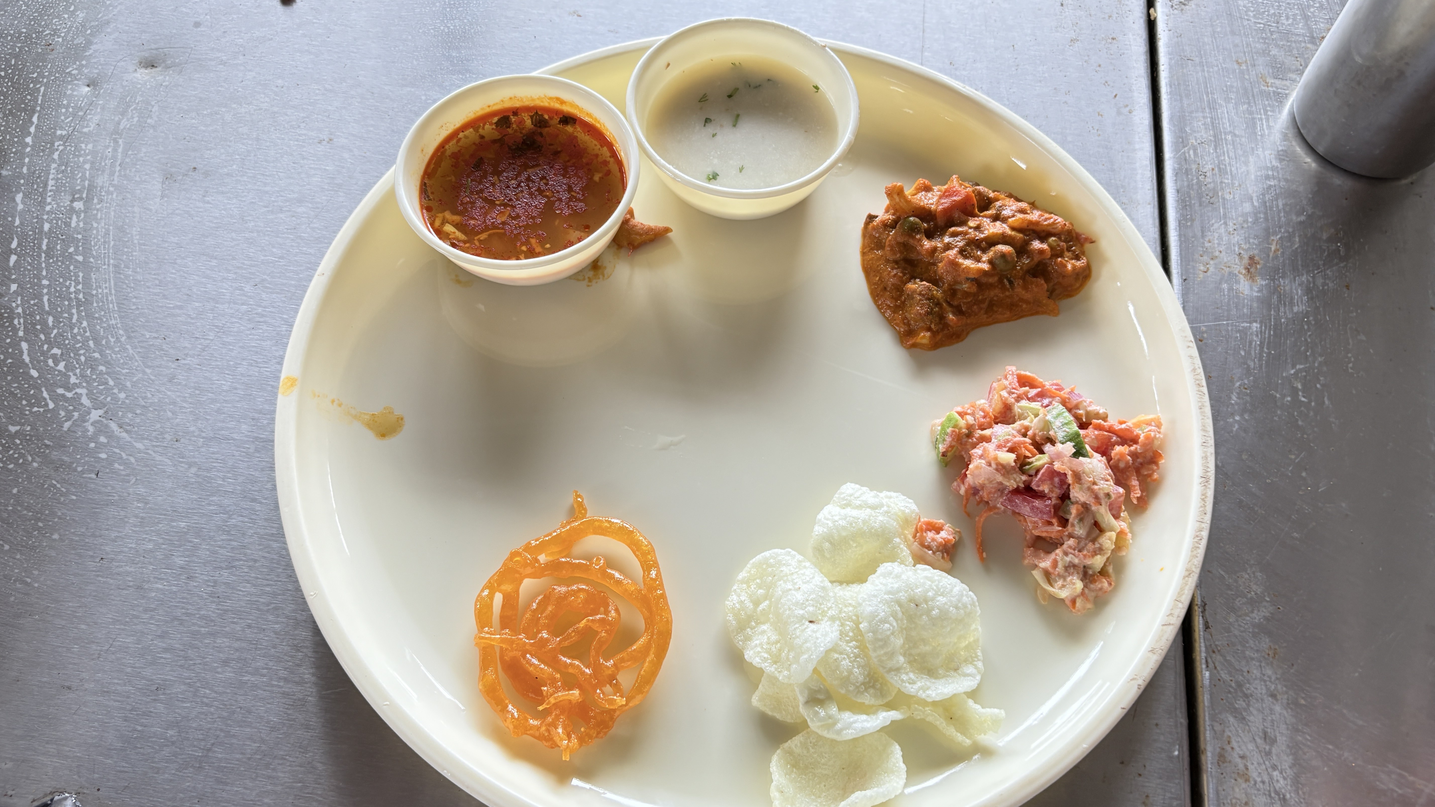 A white plate on a metal table featuring a colorful spread of Indian cuisine: red lentil dal, herbed yogurt, brown vegetable curry, crisp white papad, orange jalebi, and vibrant pink-and-white vegetable chaat.