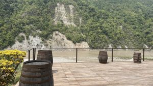 A wooden deck overlooks a river, surrounded by lush green hills and steep cliffs in the background.