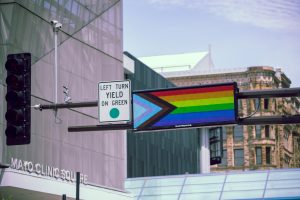 A digital traffic sign displays the Progress Pride flag above a busy city intersection. Nearby, a street sign reads “LEFT TURN YIELD ON GREEN,” and the Mayo Clinic Square building and historic architecture are visible in the background.
