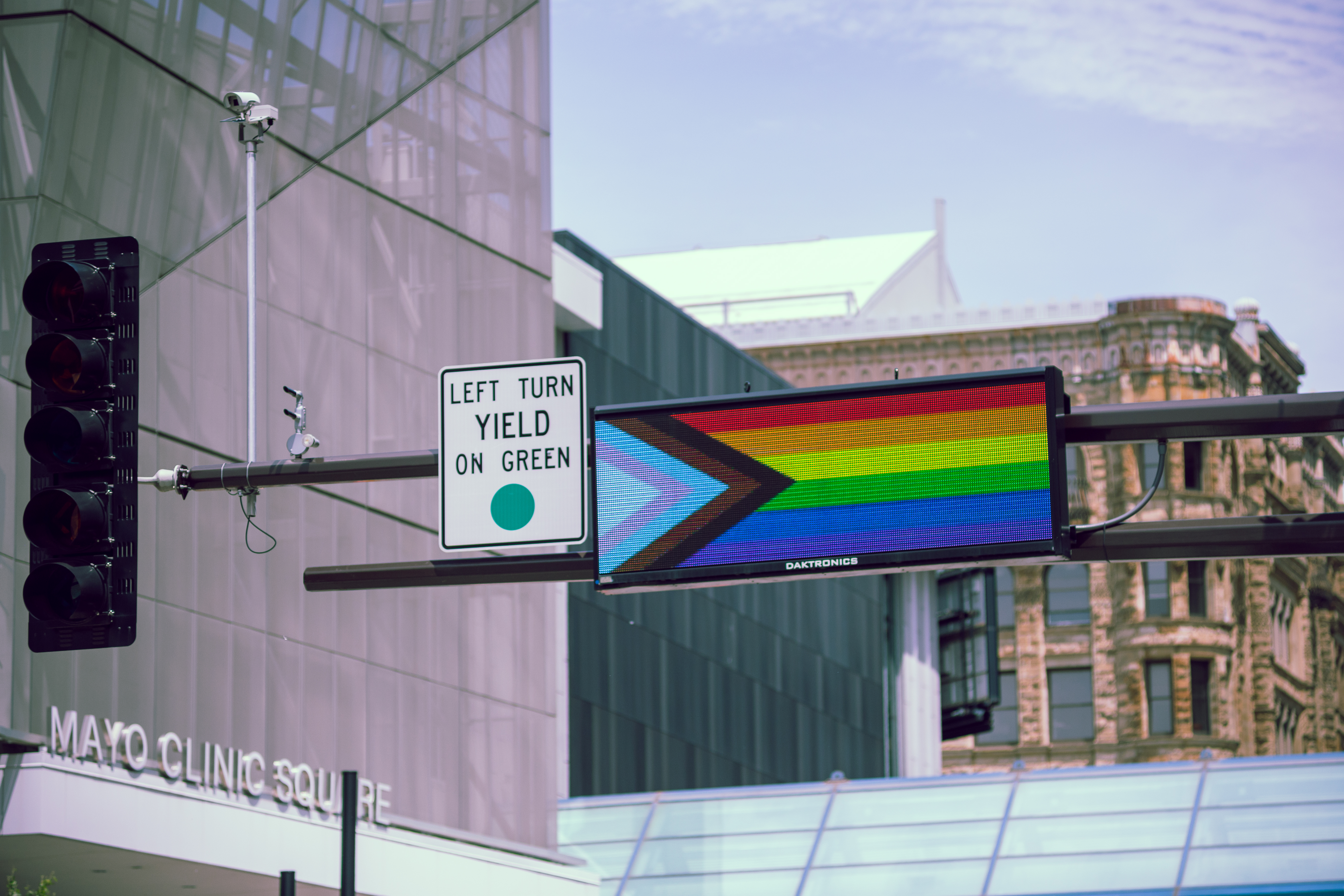 A digital traffic sign displays the Progress Pride flag above a busy city intersection. Nearby, a street sign reads “LEFT TURN YIELD ON GREEN,” and the Mayo Clinic Square building and historic architecture are visible in the background.