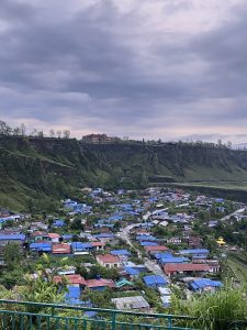 A panoramic view of a hillside village nestled in a lush valley. The village features numerous houses with blue roofs, interspersed among greenery. In the background, steep cliffs rise, with a building visible at the top. The sky is overcast with gray clouds, giving a moody atmosphere to the scene.