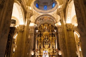 The image depicts the interior of a grand, ornate cathedral with high, arching ceilings and intricate architectural details. Dominating the scene is an elaborately carved altar adorned with golden figures and religious iconography.