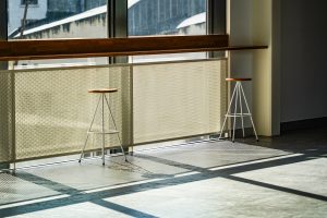 A bright indoor space featuring two minimalist stools with wooden seats and white metal legs. They are positioned near a large window with sunlight casting geometric shadows on the floor.