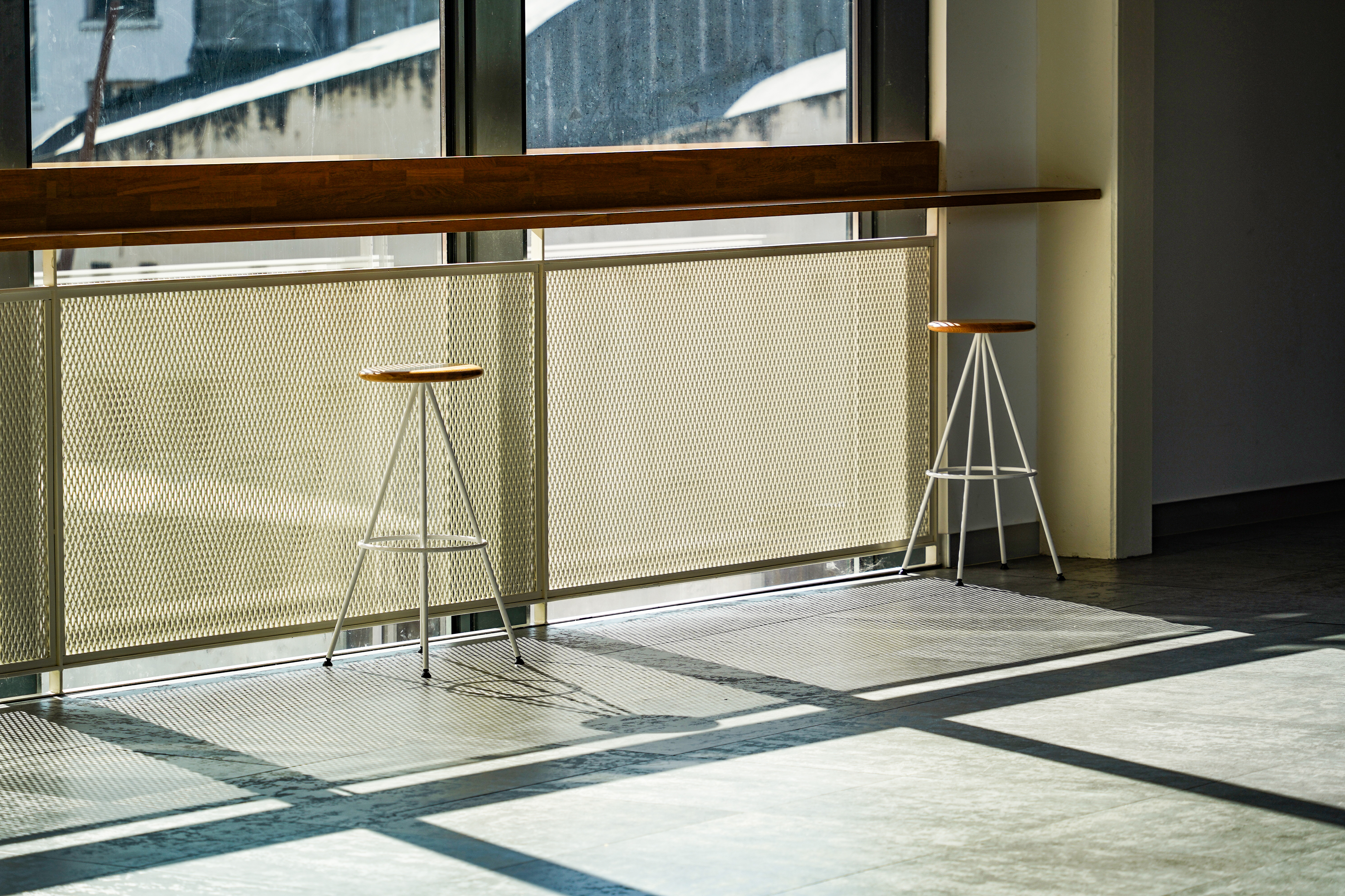 A bright indoor space featuring two minimalist stools with wooden seats and white metal legs. They are positioned near a large window with sunlight casting geometric shadows on the floor.