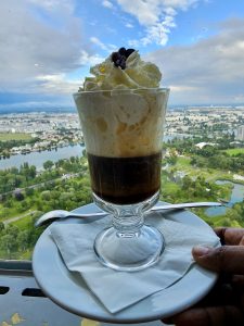 A layered coffee drink topped with whipped cream and a few chocolate-covered coffee beans, served in a glass on a plate. Photo is taken from Danube Tower, Vienna. 