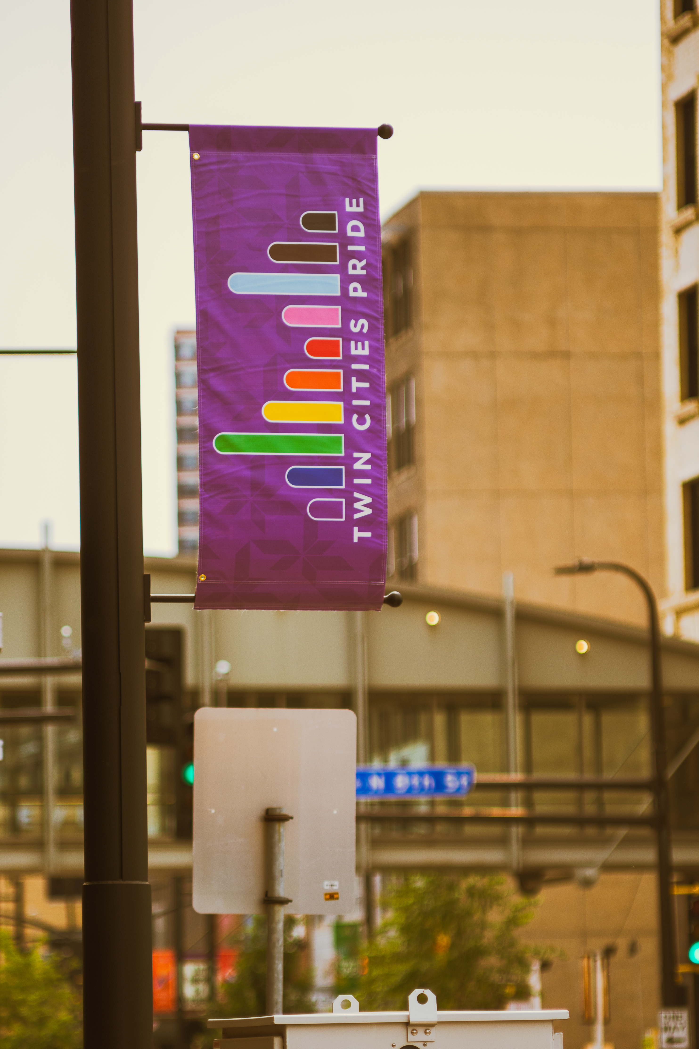 A purple banner reads "Twin Cities Pride" with a rainbow bridge logo and hangs from a light pole. In the background is a cityscape.
