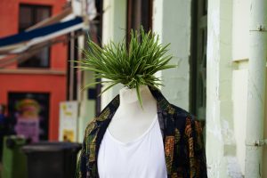 A mannequin dressed in a patterned shirt and a white tank top is standing outside. Instead of a head, it has a tuft of green grass-like plants protruding from the top. 
