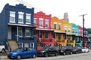 A staggered row of small city houses painted in vivid colors: dark blue, red, orange, yellow, turquoise, red, dark blue. (Address numbers have been removed with inpainting.)