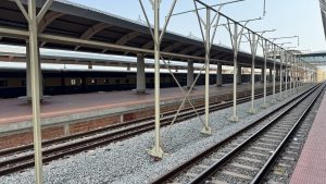 An empty railway station platform with long, metallic columns supporting the roof. In the background, blue and beige train cars are visible, and the platform is lined with brown bricks. 