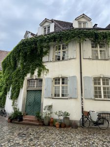 A white stucco house with green shutters, covered in ivy, features potted plants by the door and two bicycles parked on a cobblestone street.

