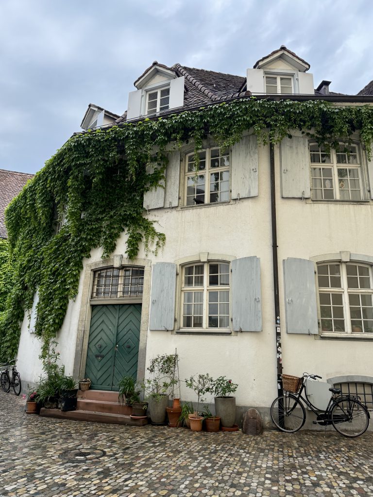 A white stucco house with green shutters, covered in ivy, features potted plants by the door and two bicycles parked on a cobblestone street.
