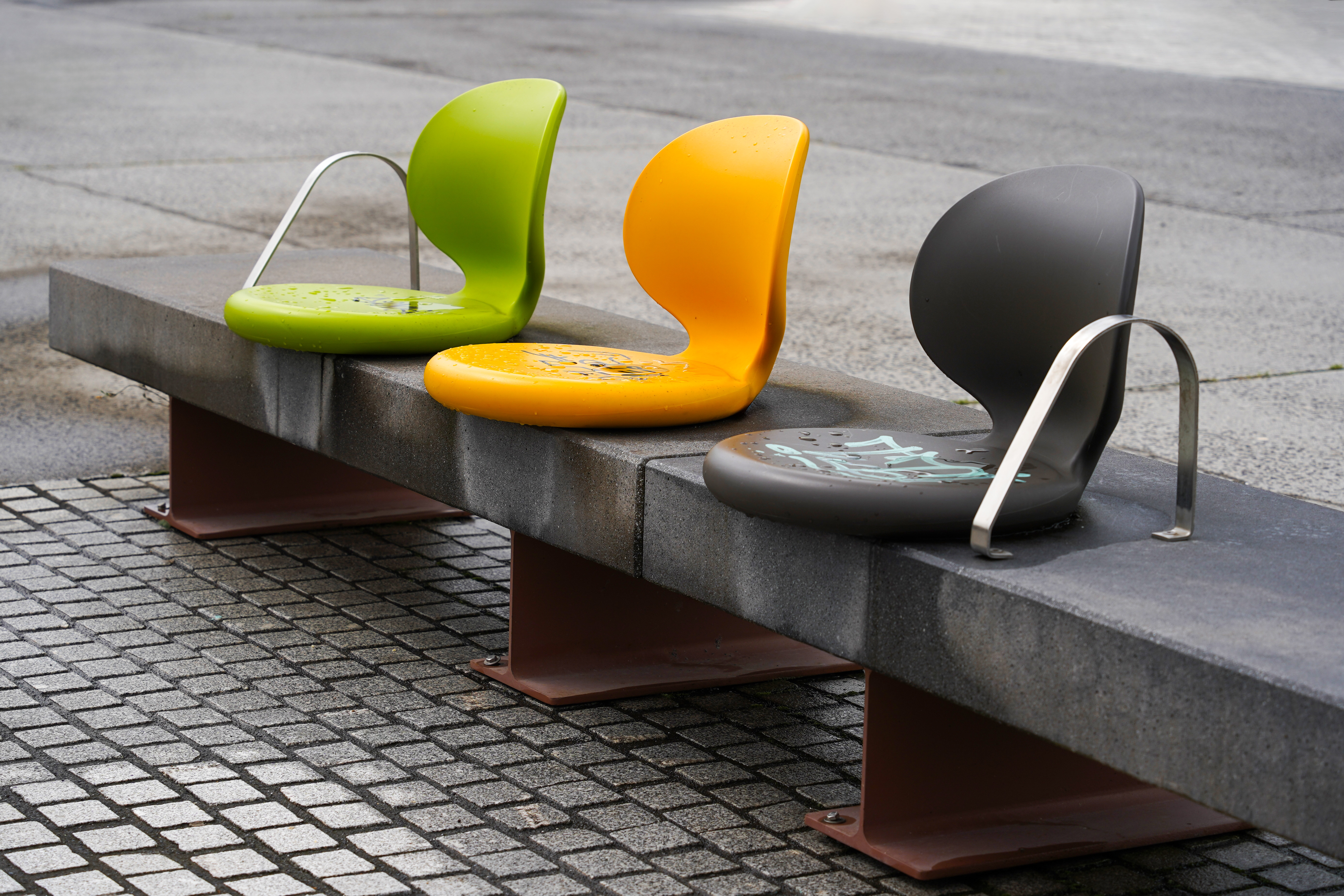A close-up image of three modern, curved seating chairs in vibrant colors: green, yellow, and dark gray. They are positioned on a gray stone bench with a rough texture, set against a backdrop of a paved area.