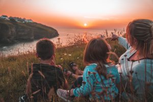 A family of three, a woman, a girl, and a boy, sit in the grass with their backs to the viewer, hugging and gazing at a vibrant sunset over a cliff and the sea. In the background, a village is silhouetted against the colorful sky.
