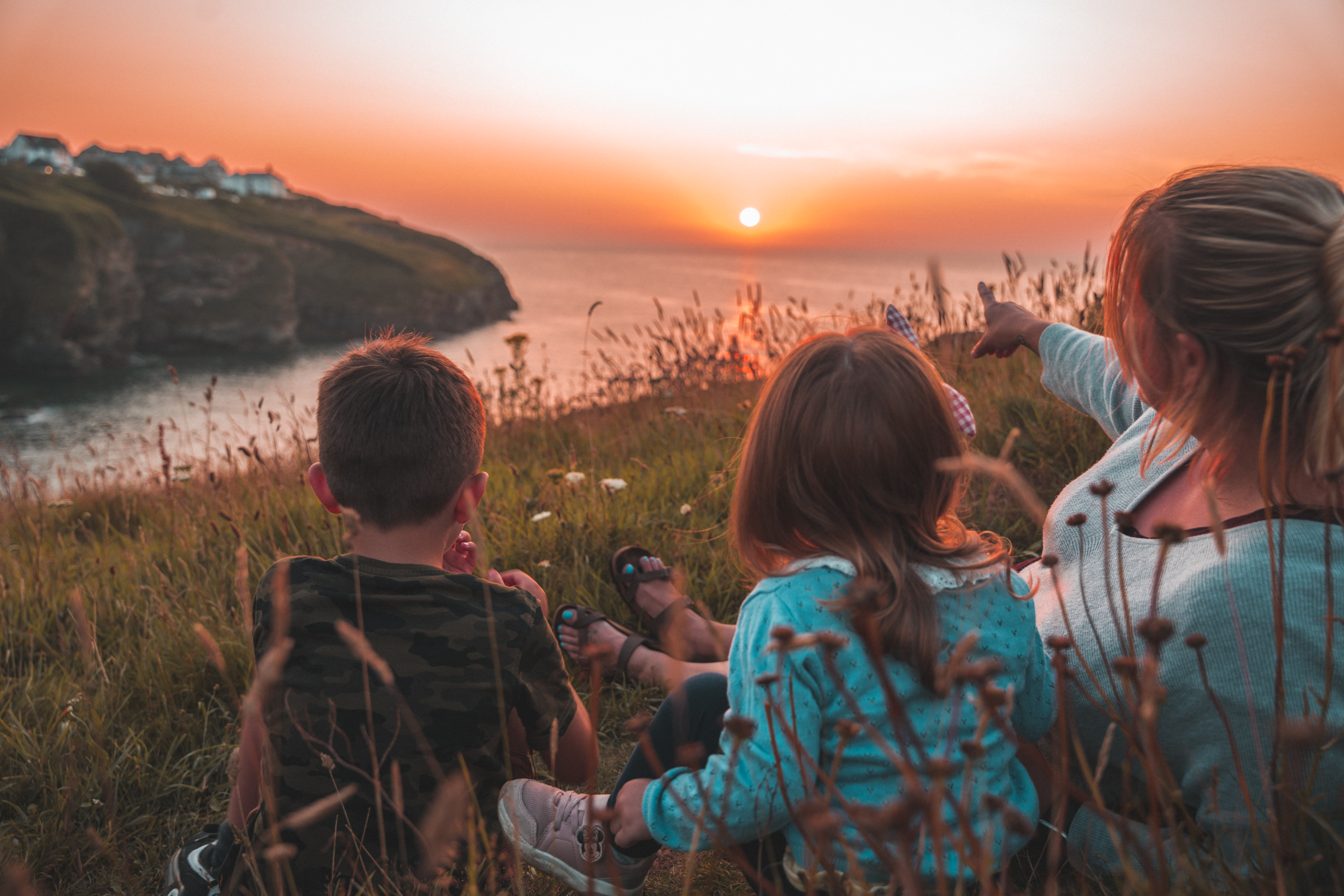 A family of three, a woman, a girl, and a boy, sit in the grass with their backs to the viewer, hugging and gazing at a vibrant sunset over a cliff and the sea. In the background, a village is silhouetted against the colorful sky.